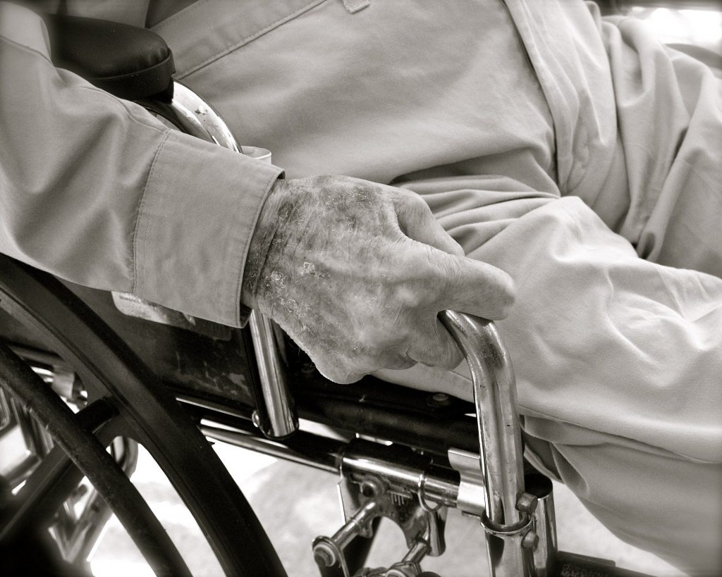 Zoom sur la main d'un personne âgé dans un fauteuil en noir et blanc qui a probablement subi de la maltraitance. 