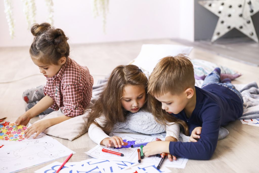 Tres niños concentrados están jugando en el suelo y dibujando en libros para colorear.